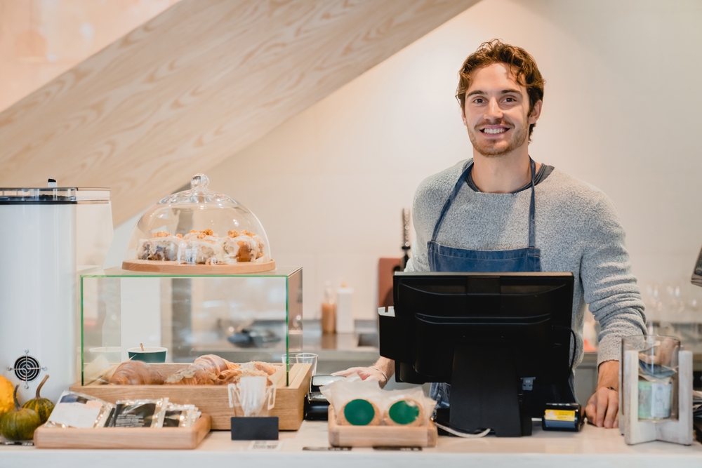 Smiling,attractive,young,waiter,standing,at,cafe,counter,in,small